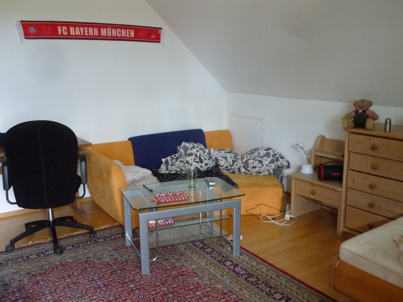 Helles Einzelzimmer im Betreuten Wohnen mit Schlafcouch, Schreibtischstuhl, Kommode und Fußballschal an der Wand